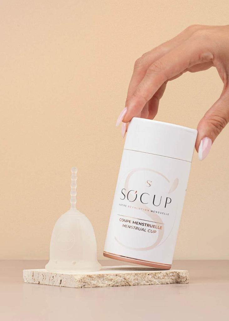 Coupe menstruelle So'Cup sur fond neutre, montrée à côté de son emballage blanc élégant. Une main féminine tient le couvercle du boîtier. Le design minimaliste et épuré du produit reflète son confort et son efficacité.